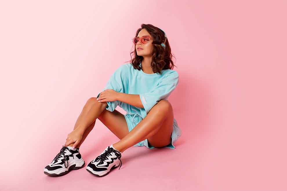 Modelo sentada com roupas azuis e óculos de sol em fundo rosa.